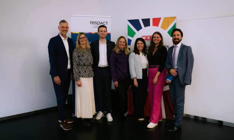 Eine Gruppe von sieben Personen steht in einem Raum vor zwei Bannern, die mit Logos und bunten Designs bedruckt sind. Sie sind elegant gekleidet und lächeln in die Kamera.