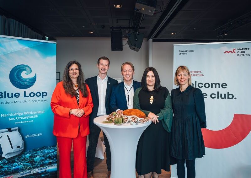 Eine Gruppe von fünf Personen steht lächelnd um einen runden Stehtisch mit Snacks. Links und rechts sind zwei Roll-Up-Banner mit den Aufschriften 'Blue Loop' und 'Welcome to the club' zu sehen.